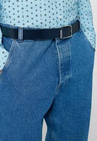 Niebieskie dżinsy z denimu o wysokim stanie i prostych nogawkach, zestawione z ciemnoniebieskim skórzanym paskiem z srebrną klamrą, noszone z wzorzystą jasnoniebieską koszulą.