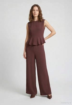 Donna che indossa un top peplum scuro senza maniche e pantaloni ampi, in piedi contro uno sfondo chiaro uniforme con una mano sui fianchi.