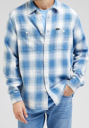 Mies, jolla on sinivalkoruudullinen nappipaita vaaleansinisen T-paidan ja sinisten farkkujen päällä, ja toinen käsi taskussa.