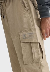 Beige cargo šortky s texturovaným povrchem, s čtvercovou náprsní kapsou na boku, nastavitelným stahovacím pasem a minimalistickými designovými prvky.