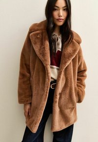 Donna che indossa un cappotto in pelliccia sintetica color camoscio sopra una camicia a righe, con lunghi capelli scuri, pantaloni scuri e una cintura nera con fibbia dorata.