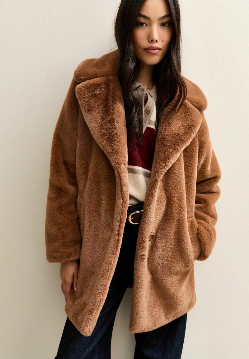 Donna che indossa un cappotto in pelliccia sintetica color camoscio sopra una camicia a righe, con lunghi capelli scuri, pantaloni scuri e una cintura nera con fibbia dorata.