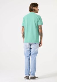 Camisa de algodón verde claro de manga corta combinada con jeans de denim azul claro y zapatillas grises, vista desde atrás. Tatuajes visibles en los brazos.