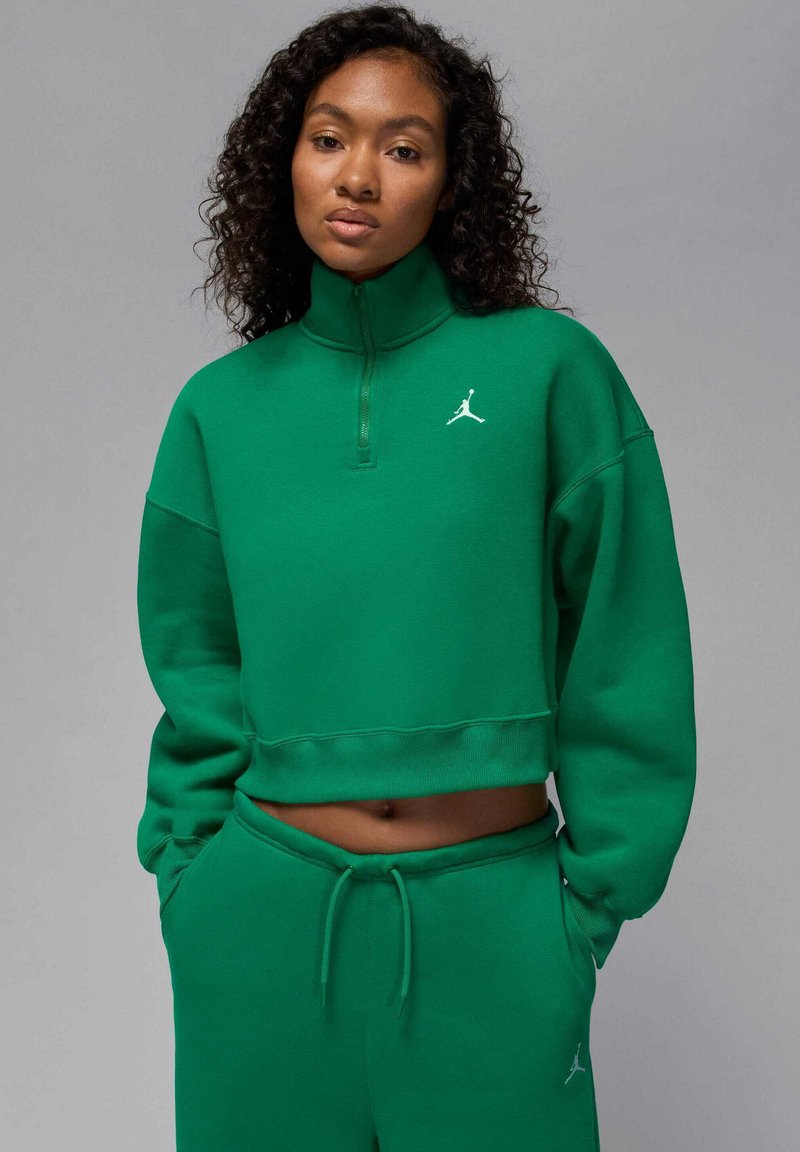 Zielona krótka bluza z wysokim kołnierzem, wykonana z miękkiego materiału, z dyskretnym białym logo na piersi, w zestawie z pasującymi spodniami dresowymi.