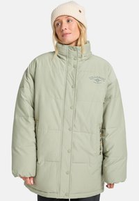 Frau trägt eine hellgrüne, übergroße gesteppte Jacke mit Druckknöpfen und einer cremefarbenen Strickmütze, schaut leicht zur Seite.