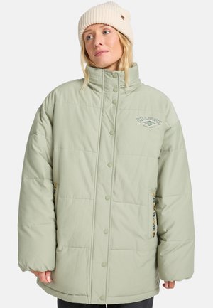 Donna che indossa una giacca trapuntata oversize verde chiaro con bottoni a scatto e un berretto di maglia crema, che guarda leggermente di lato.