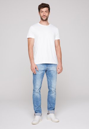 Mann steht und trägt ein schlichtes weißes T-Shirt, hellblaue Jeans mit Umschlägen und weiße Sneaker vor einem einfarbigen hellgrauen Hintergrund.