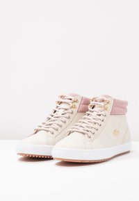 Zapatillas altas de caña con un suave cuerpo de cuero beige, cuello de punto rosa y ojales dorados. Suela blanca con patrones de tracción marrones.