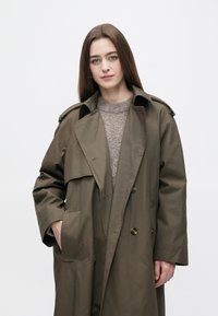 Olivgrön trenchcoat med axelklaffar, dubbelknäppt knappar och sidofickor, dragen över en beige texturerad tröja.