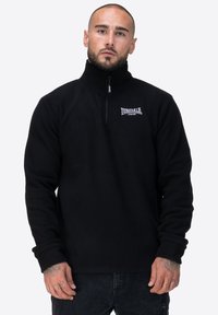 Czarny polarowy sweter z wysokim kołnierzem i zamkiem błyskawicznym na ćwierć. Posiada małe białe logo na lewej piersi. Miękka struktura, długie rękawy.