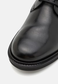 Primer plano de la parte frontal de un zapato de cuero negro pulido con costuras visibles a lo largo del borde de la suela.