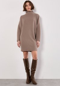 Donna che indossa un vestito a tubino beige a coste con collo alto e stivali di pelle marroni fino al ginocchio, in piedi contro un muro bianco semplice.
