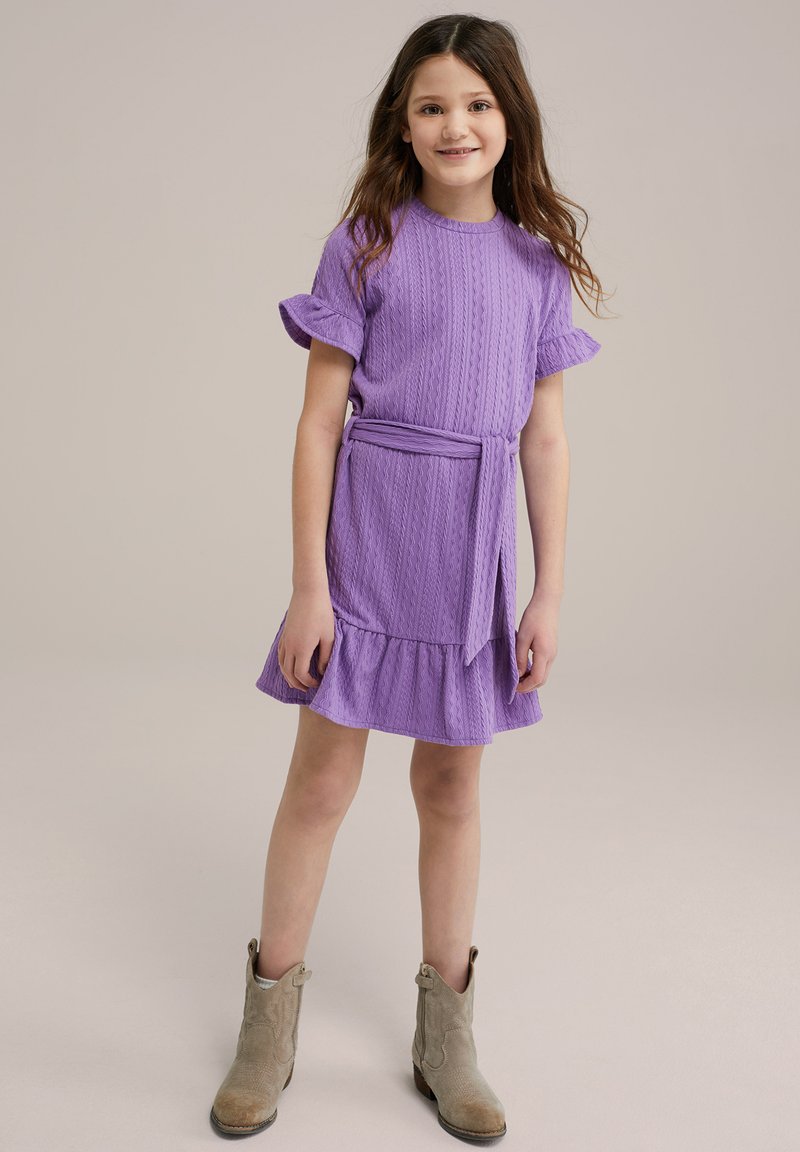 Violetinės spalvos suknelė su trumpomis volanėlėmis, tekstūruota raštas, diržas ties juosmeniu ir keliais sluoksniais volanos. Pora su šviesiai rudais kulkšnies batais.
