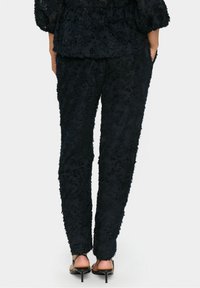 Osoba stojąca z rękami w kieszeniach, ubrana w teksturowane czarne spodnie i pasującą górę, w zestawie z czarnymi sandałami na obcasie z otwartym tyłem.