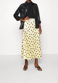 Vrouw in een zwarte leren jack, zwarte top, gele satijnen midijurk met zwarte stippen en zwarte high heels staat voor een effen achtergrond.