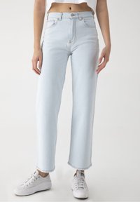 Jeans de talle alto en azul claro con corte recto, que cuentan con cinco bolsillos, costuras en contraste y cierre de botón. Combinados con zapatillas blancas.