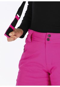 Fuchsia sneeuwbroek met een hoge tailleband, twee zwarte knoopsluitingen, een zijzak met rits en een verstelbare zwarte band in de hand.