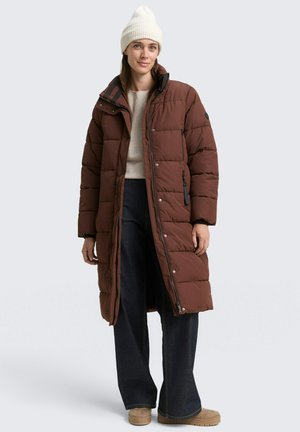 Bruine gewatteerde lange jas met een ritssluiting aan de voorkant, knoopsluitingen, zijzakken en een hoge kraag; gedragen over een beige sweater en brede jeans.