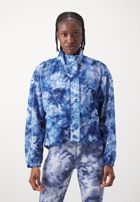 Modrá tie-dye bunda s vysokým límcem a elastickými manžetami. Obsahuje malé zelené logo Lacoste. V páru s odpovídajícími legínami se vzorem.
