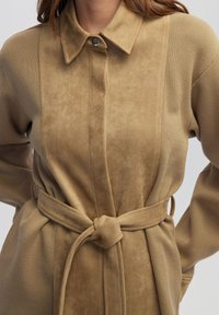 Chaqueta de ante sintético beige con cuello tipo solapa, cierre de botones y un cinturón atado en la cintura, con una textura sutil y tela suave.