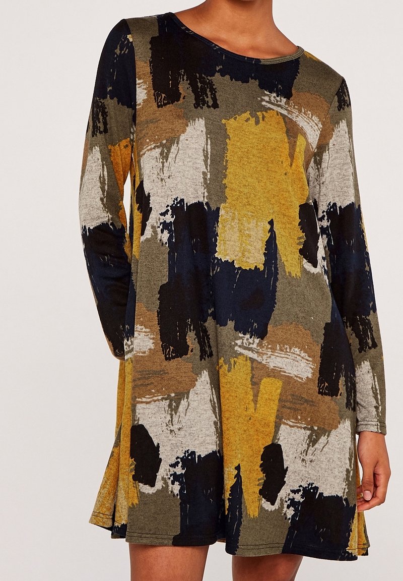 Femme portant une robe à manches longues avec un imprimé abstrait effet coups de pinceau dans des tons jaune moutarde, noir, blanc, marron et vert olive.