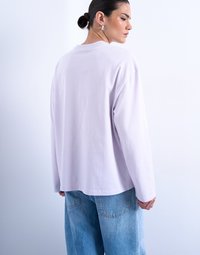 Γυναίκα με σκούρα μαλλιά σε κότσο, που φοράει ασημένια σκουλαρίκια κρίκους, μια άνετη ανοιχτό μωβ μπλούζα με μακριά μανίκια και μπλε τζιν, μπροστά από λευκό φόντο.