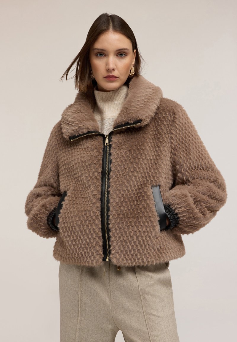 Giacca in ecopelliccia marrone con chiusura a zip, ampio colletto e motivo testurizzato. Dettagli neri sui polsini e sulle tasche frontali.