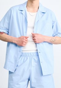 Světlomodrá košile s krátkým rukávem na knoflíky s náprsní kapsou, doplněná o shodné kraťasy. Pod tím bílý tílko. Texturovaná látka.