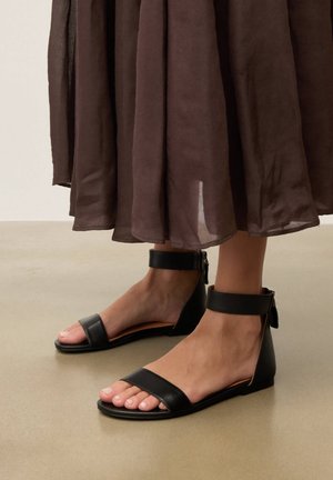 Jalat mustat matalat sandaalit nilkkaremmeillä, yhdistettynä ruskeaan puoliläpinäkyvään liehuvaan hameeseen, seisomassa beigenvärisellä lattialla.