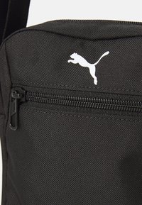 Bolso bandolera de tela negra con una superficie texturizada, que cuenta con un bolsillo con cremallera y un logo de Puma en blanco. Incluye una correa ajustable.
