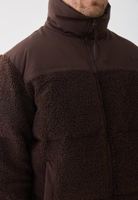 Brun fleecejakke med høy krage, teksturert kropp og glatte ermer. Har en frontglidelås og brodert logo på brystet.