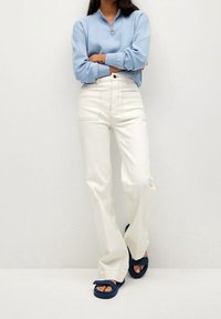 Jersey azul claro con cremallera en el cuello, jeans blancos de pierna ancha con bolsillos delanteros y sandalias azul marino. Texturas suaves y detalles de costuras en contraste.