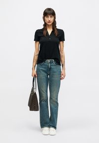 Mulher com cabelo comprido e escuro, vestindo uma polo preta, jeans azuis, ténis brancos, segurando uma bolsa com padrão, a posar contra um fundo claro e liso.