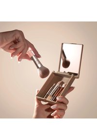 Hånd holder sminkekost over et åpent etui med fire andre koster og et speil som reflekterer kosten.