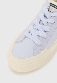 Light lavender canvas sneaker with flat laces, cream rubber sole, and stitched accents. Features a rounded toe and textured upper.