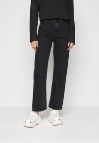 Vaqueros negros de cintura alta con corte recto y un ligero desgastado, combinados con zuecos blancos que tienen agujeros de ventilación y una suela gruesa.