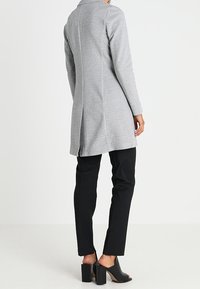 Manteau gris à manches longues avec col châle, coupe droite et fente arrière. Associé à un pantalon noir et des sandales à talons ouvertes.