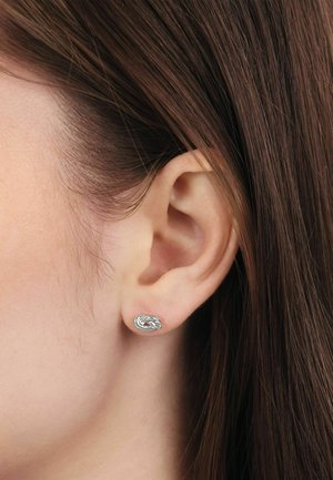 Primo piano dell’orecchio di una persona con capelli castani e lisci, che indossa un piccolo orecchino a bottone d’argento a forma di nodo intrecciato.