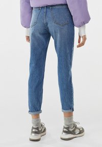 Džínové kalhoty s vysokým pasem v modré barvě, s tapered nohavicemi, ohrnutými manžetami a zadními kapsami. Nosí se s šedými pletenými ponožkami a šedými teniskami.