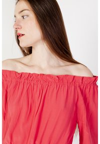 Top corallo off-the-shoulder con scollo arricciato, tessuto fluido, texture morbida e drappeggio leggero. Presentato su uno sfondo semplice.