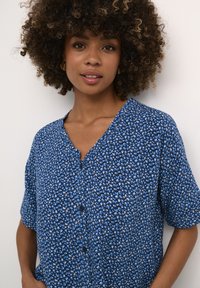 Mėlyna gėlių trumpomis rankovėmis marškinėliai su V formos iškirpte ir mygtukų užsegimu. Audinys turi švelnų paviršių, su sudėtingais šviesiai mėlynais žiedais tamsiame fone.