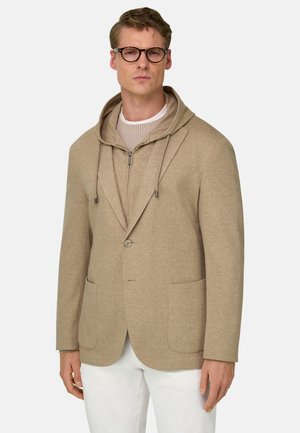Man draagt een beige hoodie met rits onder een bijpassend beige colbert, een witte trui, witte broek en ronde zwarte bril, kijkt recht vooruit.
