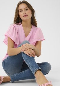 Svetlo roza majica s V-izrezom in slojevitimi volanastimi rokavi ter čipkastimi detajli, v kombinaciji s oprijetimi modrimi džinsami in svetlo roza čipkastimi čevlji na vezanje.