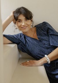 Žena s tmavými vlasmi v tmavomodrých šatách s bodkami a striebornými šperkami, opierajúca sa o béžovú pohovku, jemne sa usmieva.