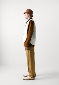 Brauner Bucket Hat, weißes Hemd über braunem Langarmshirt, beige Hosen mit Taschen, weiße Sneakers mit bunten Stickdetails.