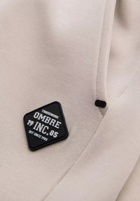 Béžová tkanina s hladkým povrchem s černě šitým štítkem s nápisem "OMBRE 19 INC. 85" bílým písmem, připevněným pod úhlem.