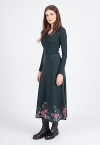 Donkergroene geribde jurk met lange mouwen, met een bloemenzoom in roze en geel. Gemaakt in combinatie met zwarte enkellaarsjes.