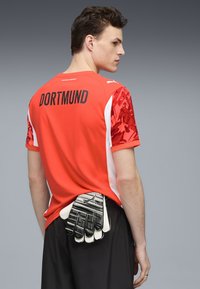 Maglia rossa e bianca con "DORTMUND" stampato sulla schiena, contrastata da maniche decorate. Guanti da portiere tenuti in vita.