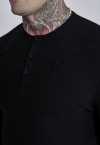 Must mustaga ribikangast valmistatud must Henley särk, millel on kolmest nuppudest koosnev nööpplacket ja väike tikandlogoga süda.