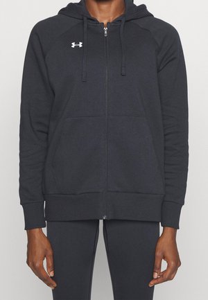 Person iført en sort lynlåshoodie med et lille hvidt Under Armour-logo på brystet og matchende sorte bukser mod en ensfarvet baggrund.